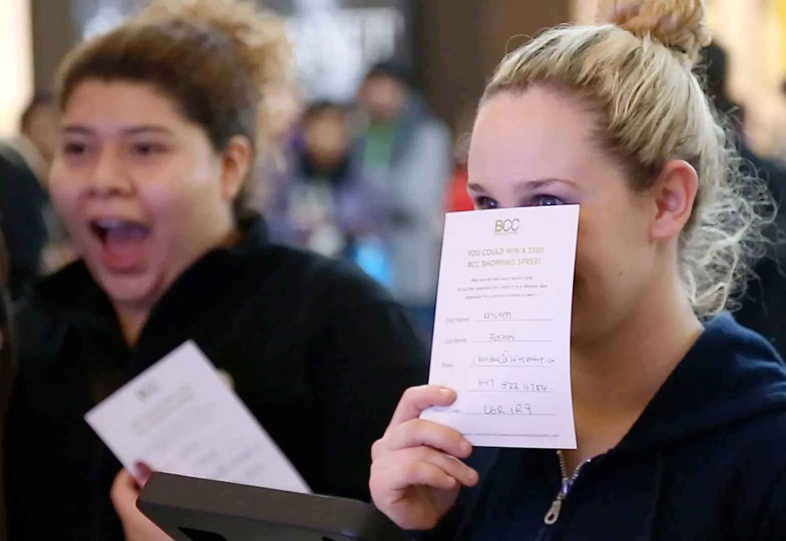 Close-up photograph of a young woman hiding her smile behind a postcard, looking with anticipation at something we can't see